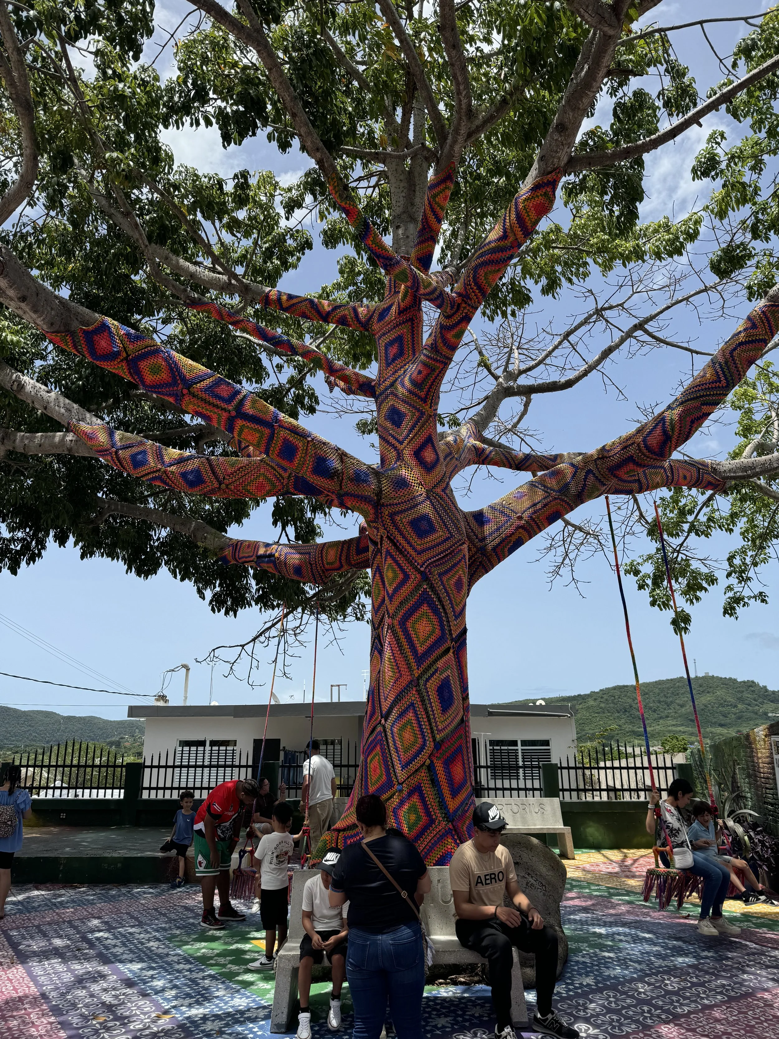 Colorful yarn-wrapped tree in a public area with children and adults, some sitting on a bench, under a partly cloudy sky near a white building with black fencing.