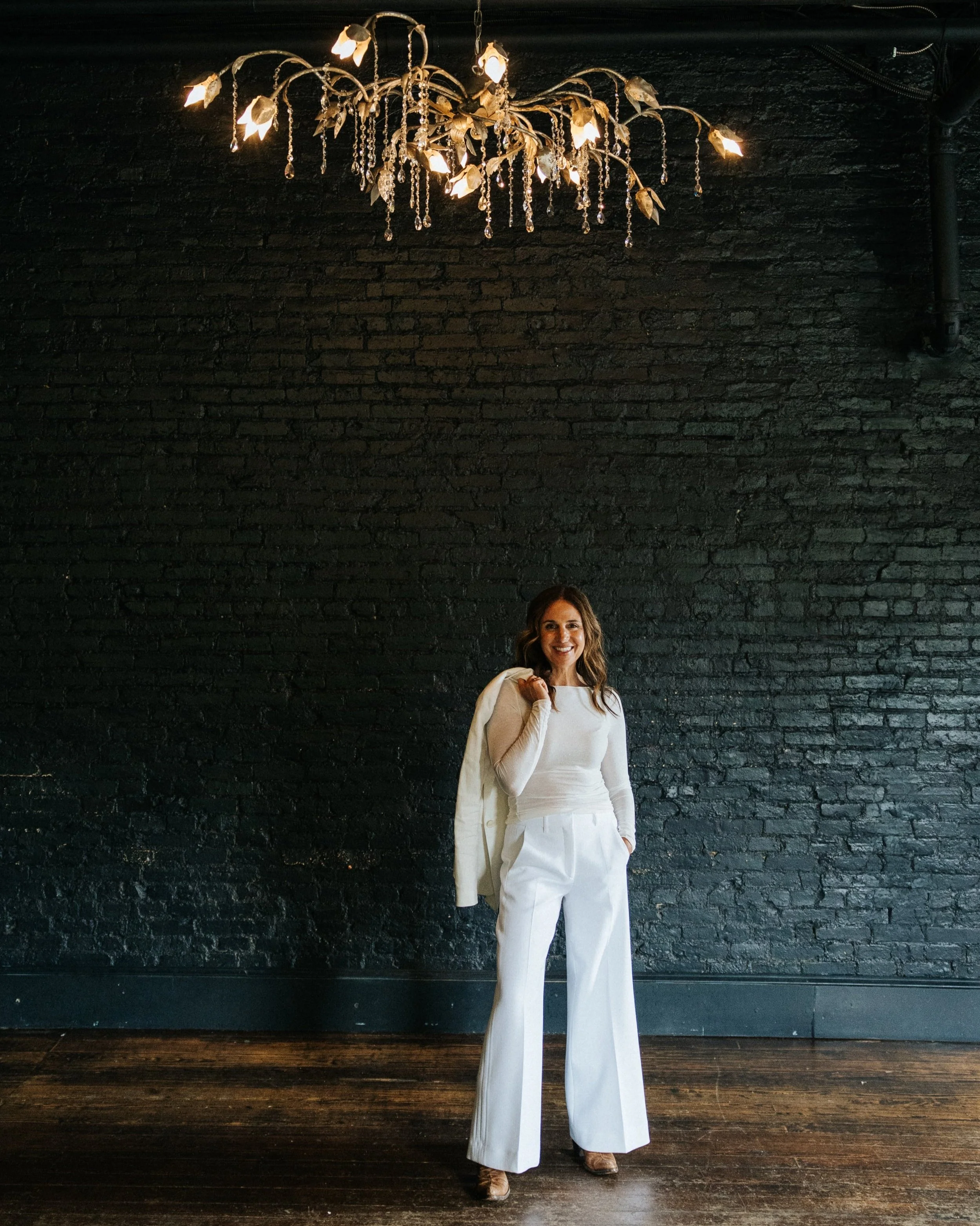 Woman in white outfit posing in front of a black brick wall with a decorative chandelier hanging above.