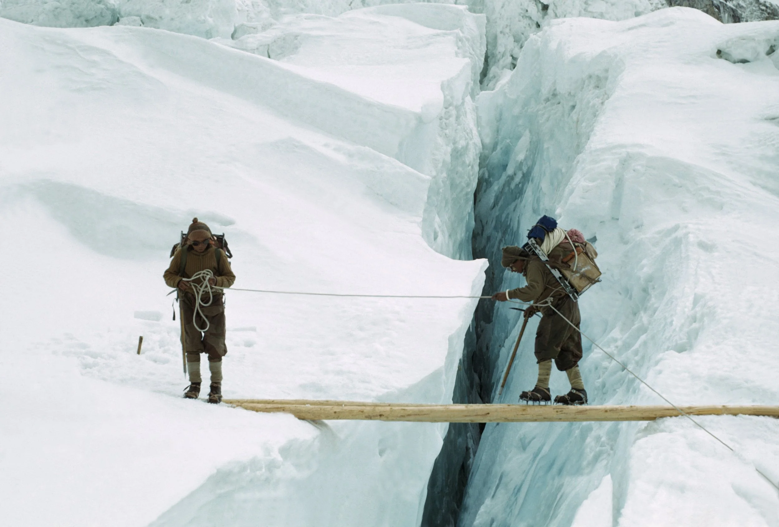 Two climbers crossing a crevasse on a wooden plank bridge in a snowy, glacial landscape.