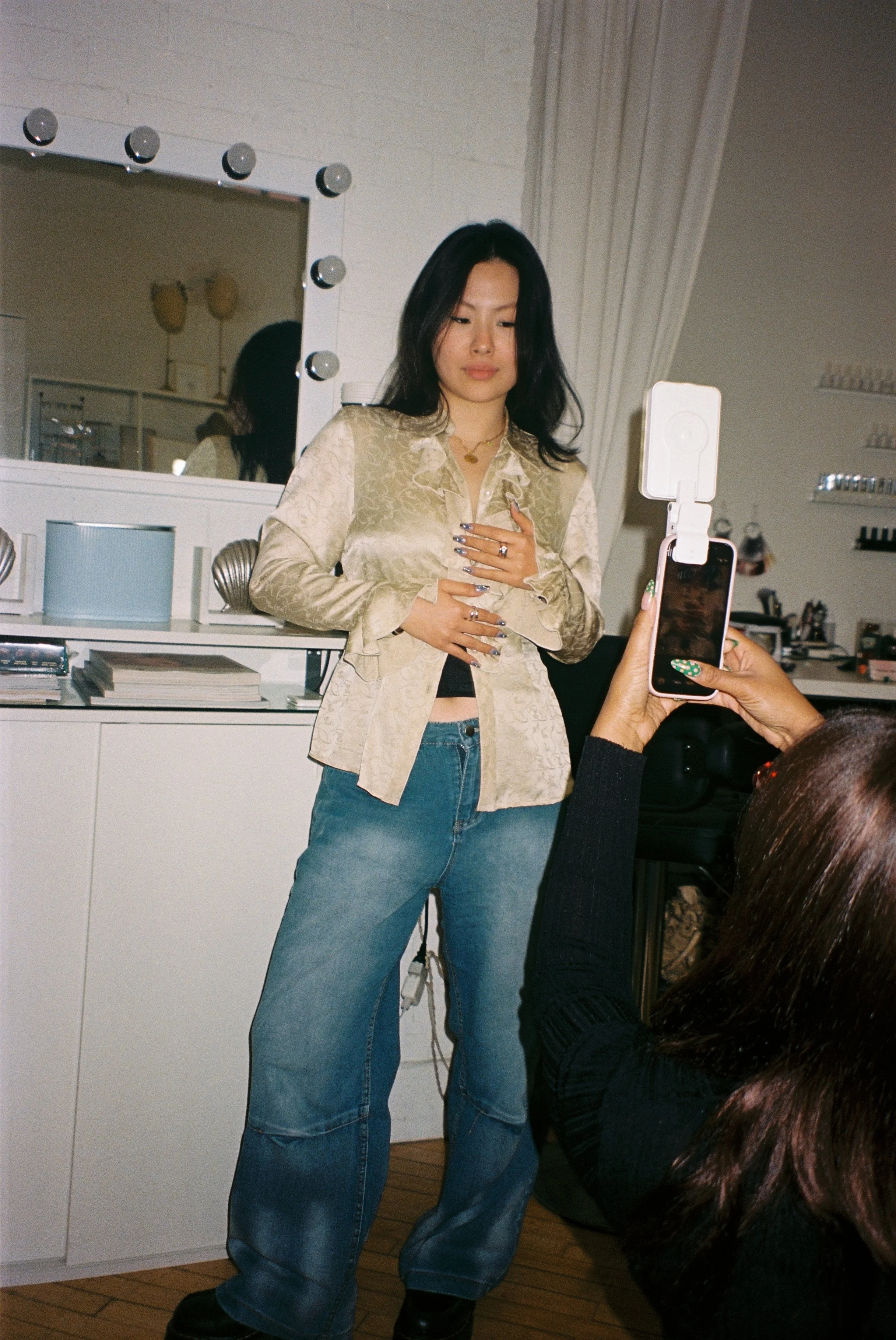 A woman with black hair wearing a beige silk blouse, blue jeans, and black boots is looking at herself in a mirror with light bulbs around the frame. She is holding her chest with one hand while a person with dark curly hair, wearing a black sweater, is taking her photo with a smartphone. The room has white walls and shelves with small items, and a wooden floor.