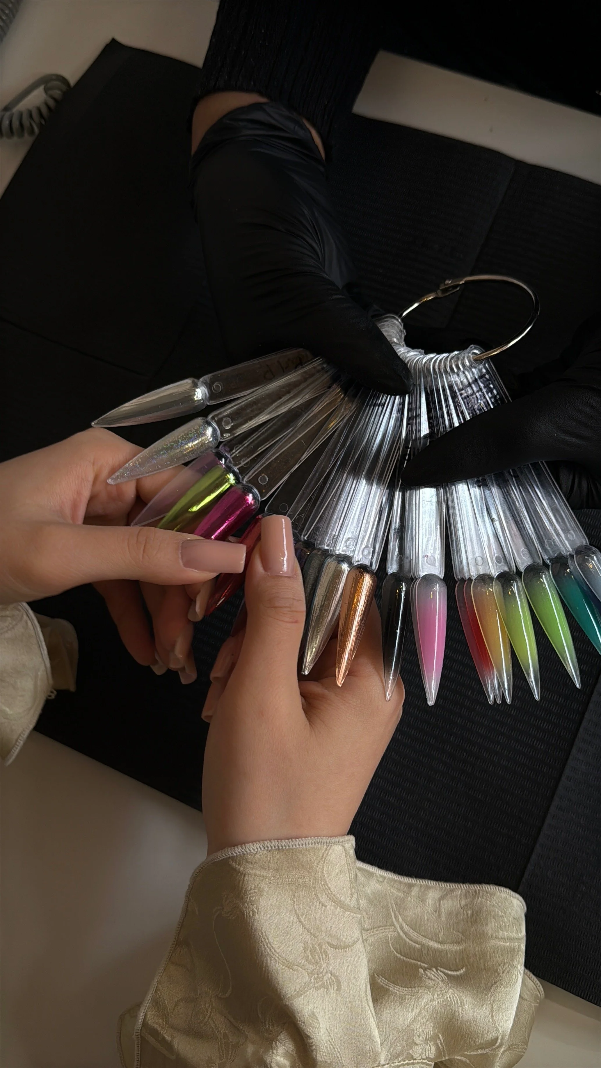 Person holding swatch of false nail tips with various colors and styles, sitting on black furniture, wearing a light-colored dress with floral pattern.