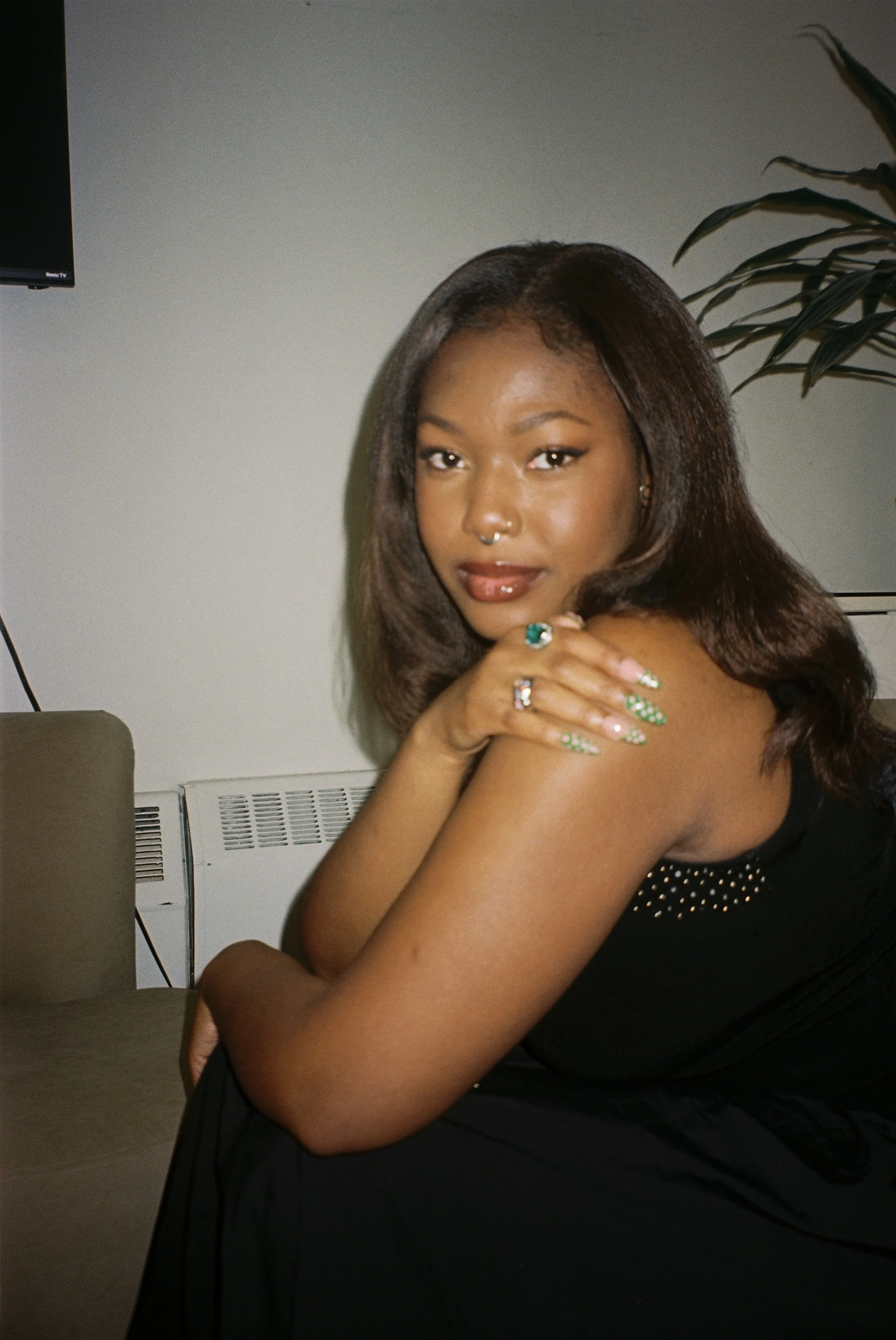 A young black woman with dark brown hair and a septum piercing, wearing a black dress with silver dots, sitting with one arm crossed and resting on a table in an indoor setting.