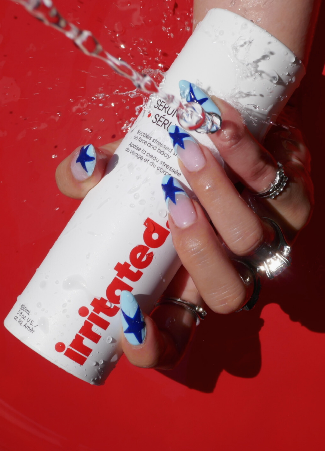 A hand with multi-ring jewelry and nails painted with white base and blue star designs is holding a white bottle of skincare product labeled 'Retise' while water is poured over it, against a red background.