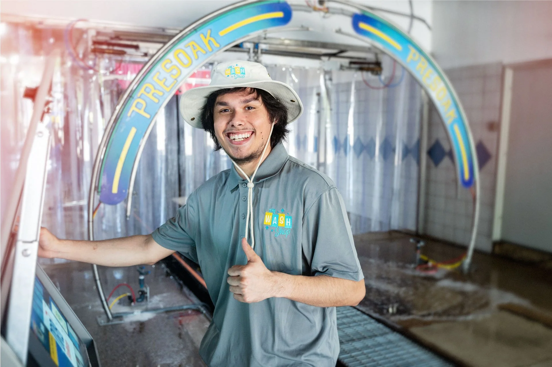 A friendly Wash Shop employee wearing a branded hat and polo shirt, smiling and giving a thumbs up inside the car wash tunnel.