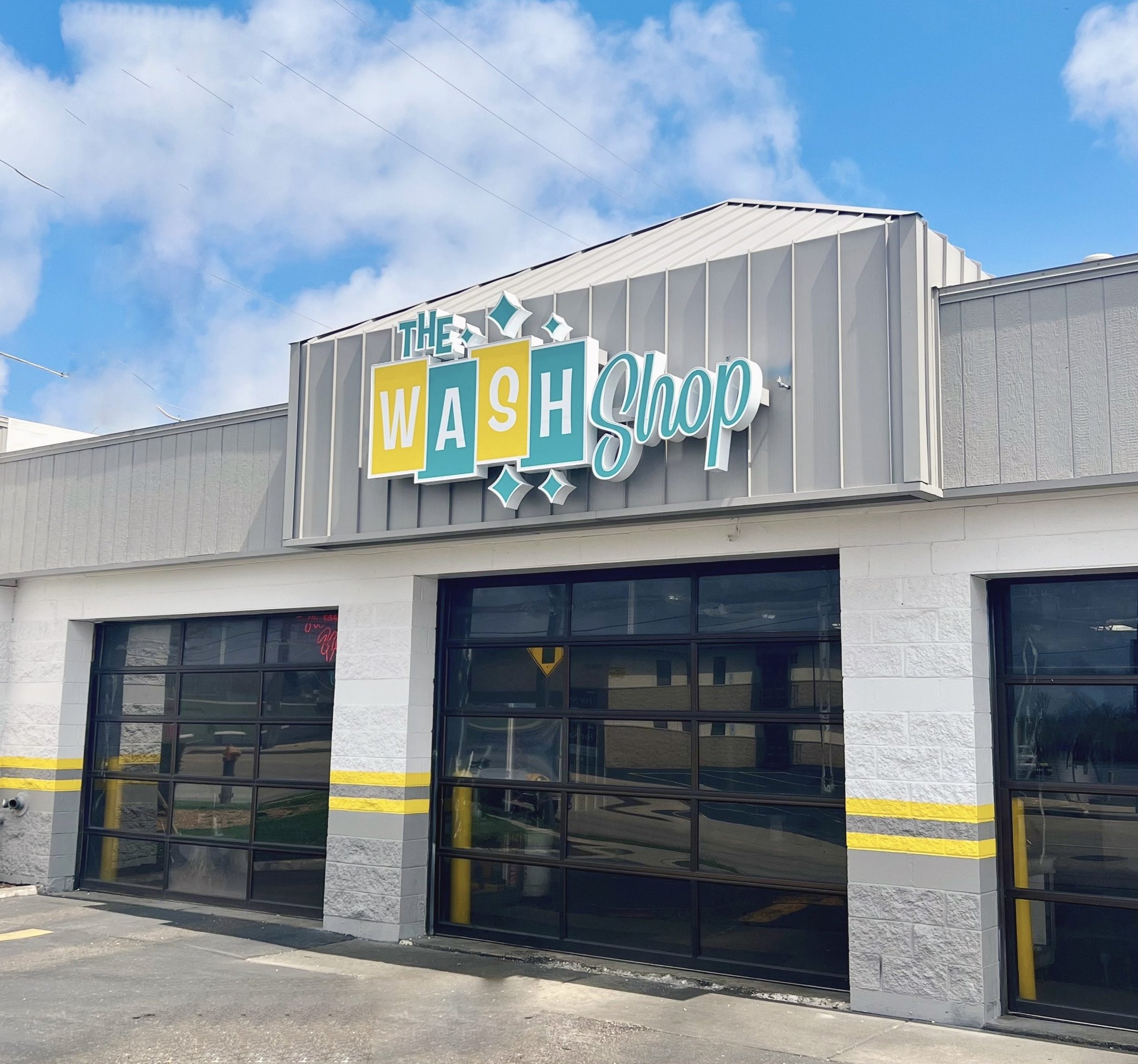 The exterior of The Wash Shop building on a sunny day. A large, retro-styled teal and yellow sign is mounted above three black glass garage doors on a white block building with a yellow painted stripe.