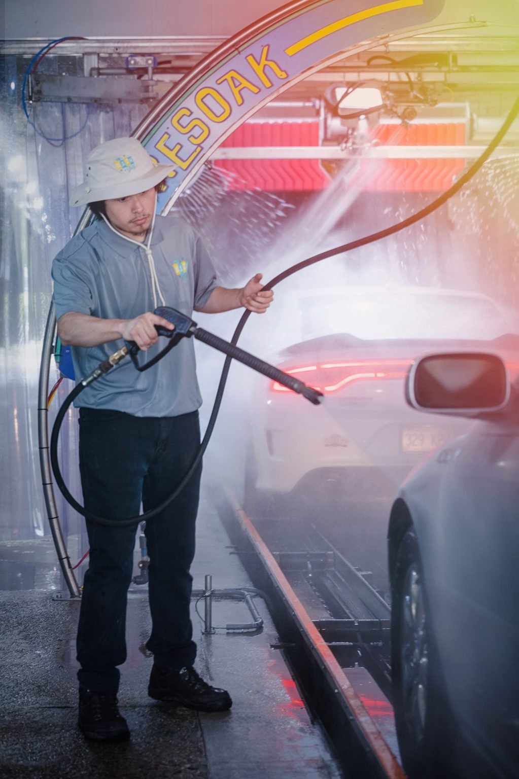 The Wash Shop attendant pressure washing the rear of a white vehicle under the illuminated presoak arch.