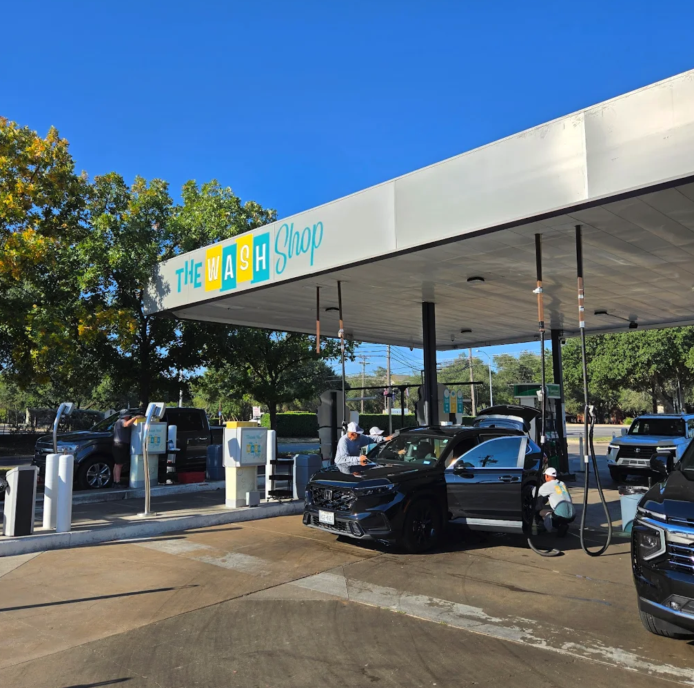 The Wash Shop employees providing full service interior detailing and vacuuming for a black Honda SUV under the shaded canopy.