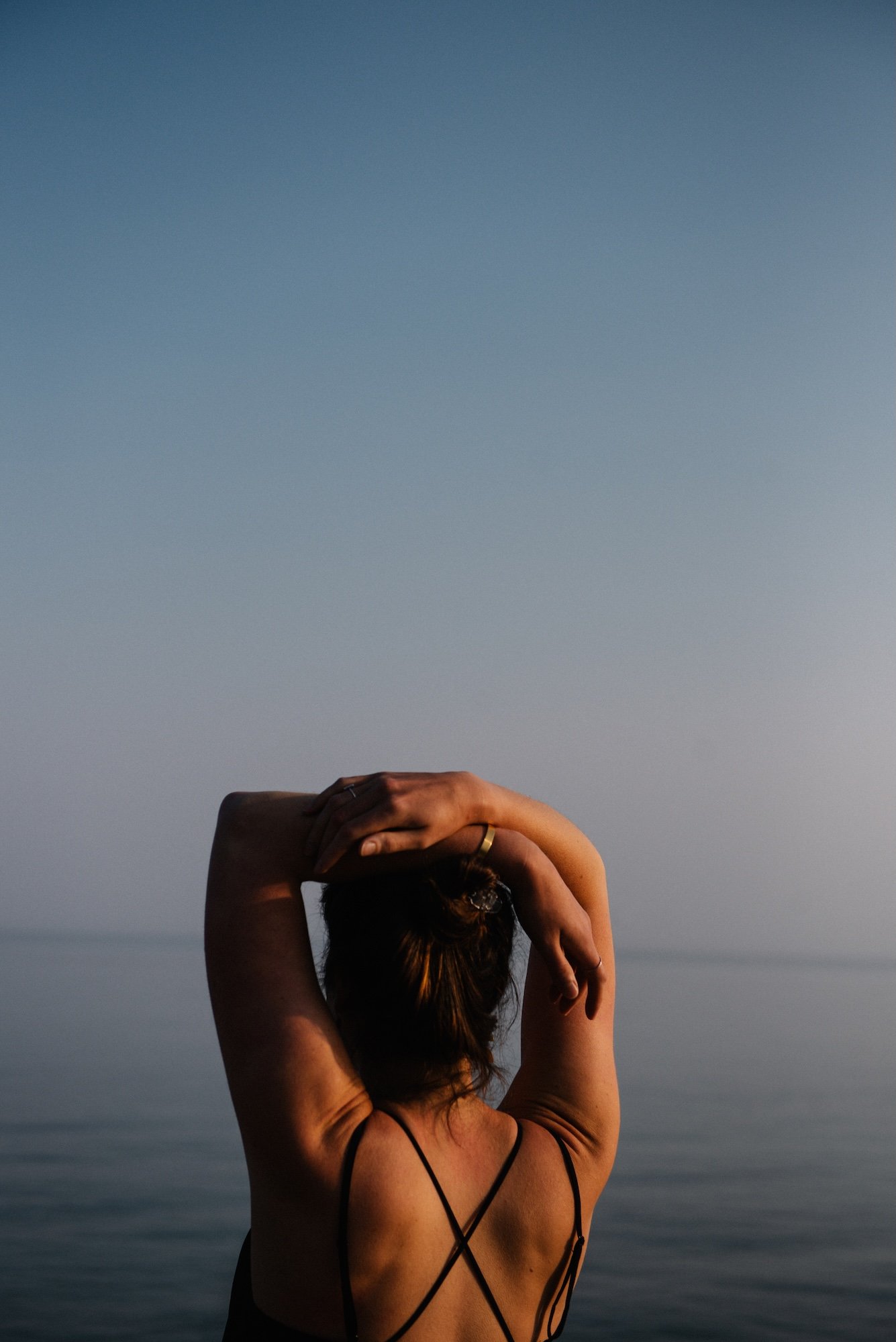 A woman with her back to the camera, arms raised and hands resting on her head, stands by a body of water during sunset or sunrise.