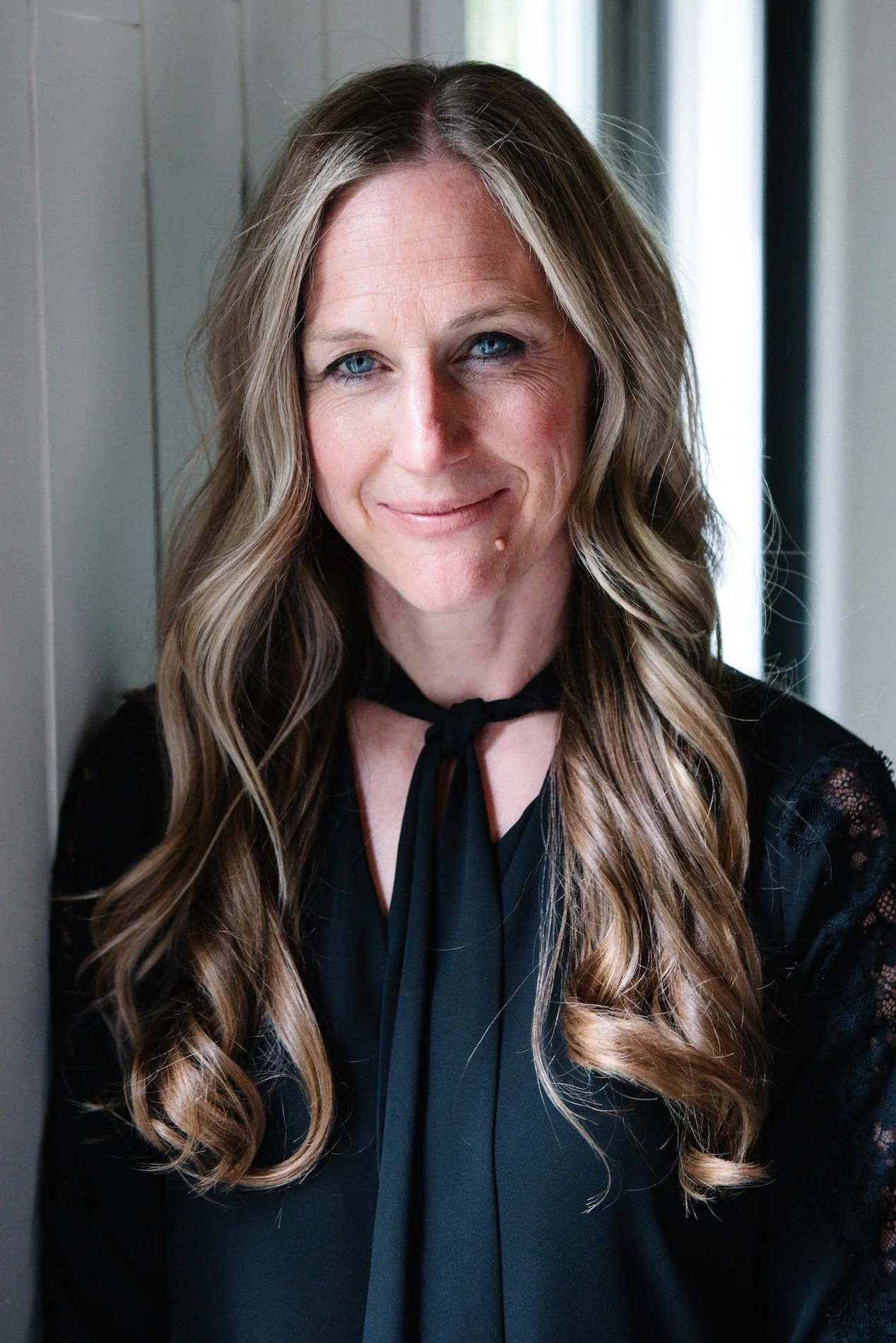 A woman with long wavy blonde hair and blue eyes, smiling softly, wearing a black top with lace details and a black scarf tied around her neck, standing indoors near a wall and window.