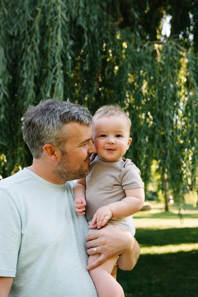 Father holding smiling baby boy outdoors with green trees in background.