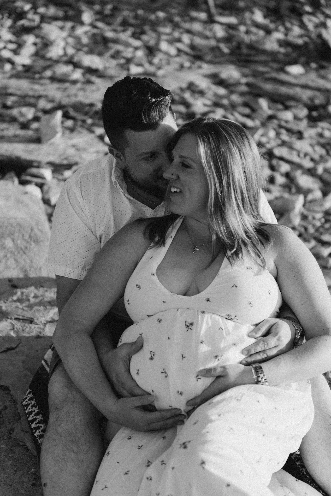 A couple sitting on a rocky beach, with the man embracing the pregnant woman from behind, touching her belly, as they share an intimate moment.