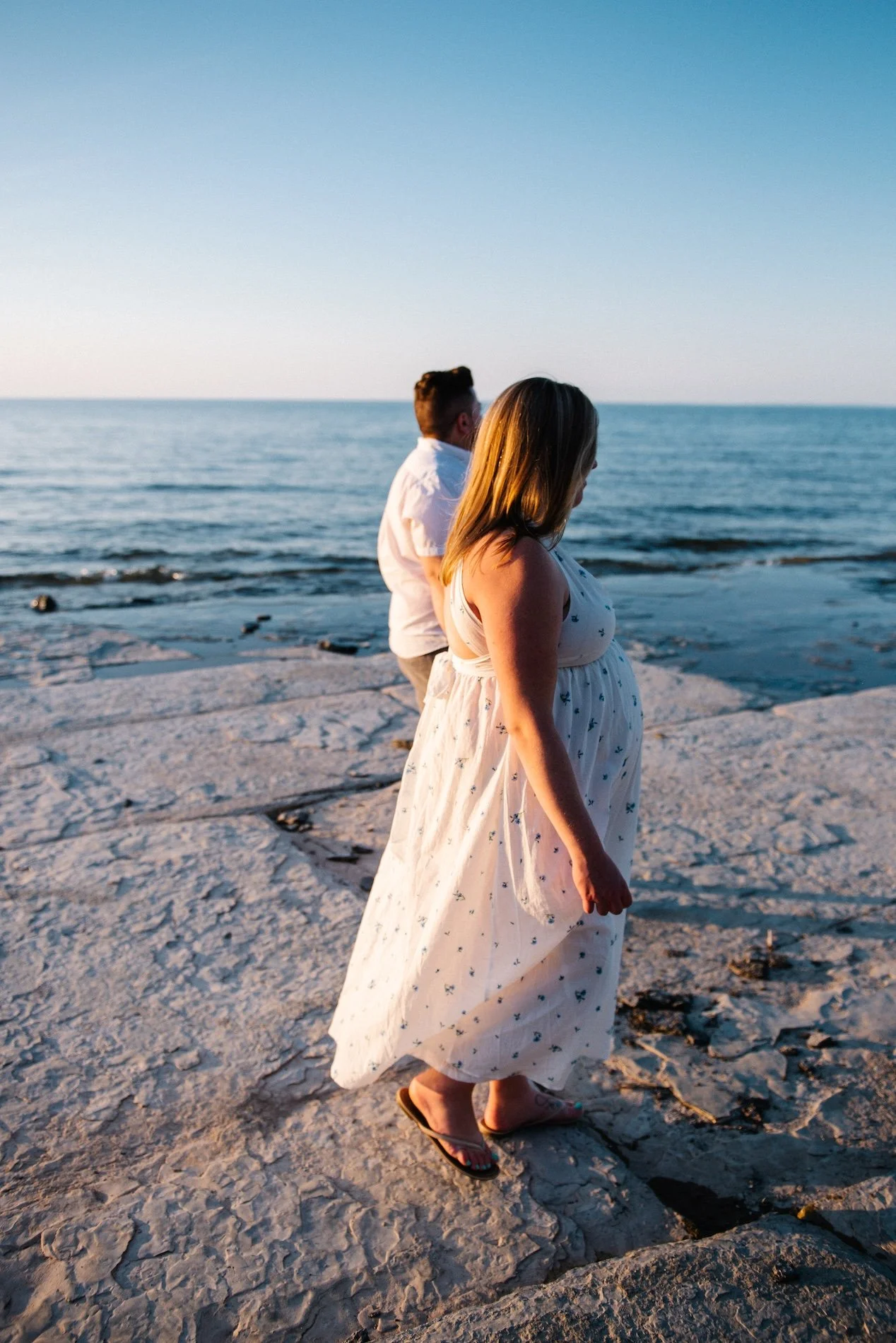 A pregnant woman in a white dress and flip-flops standing on a rocky shoreline, with a man in a white shirt and gray pants standing behind her, looking out at the ocean during sunset.