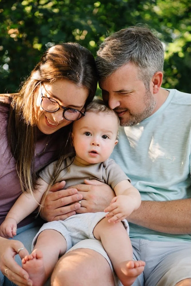 A young family of three sitting outdoors on a sunny day, with a woman, a man, and a baby boy, all close together and smiling.