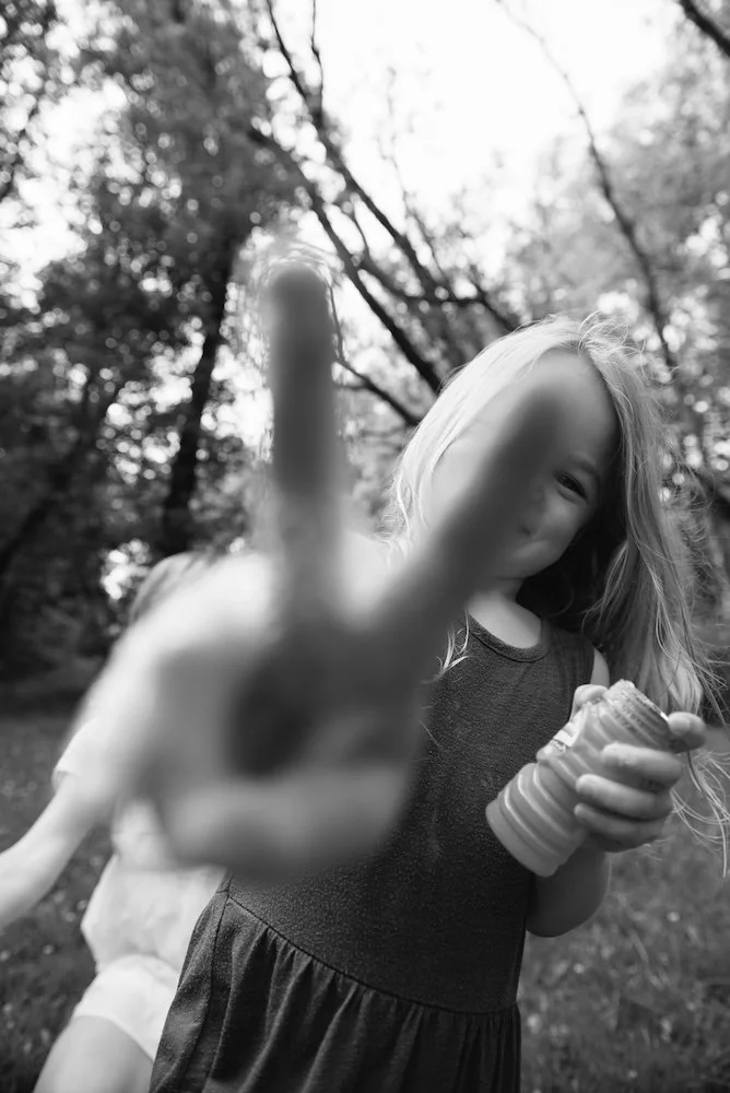 A young girl with long hair in a sleeveless dress making a peace sign with her fingers towards the camera, holding a cup or container, with a girl in light-colored clothing behind her outdoors in a wooded area.