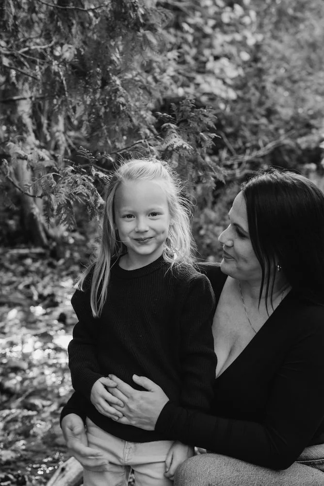 A woman with dark hair and a young girl with blonde hair, both smiling, outdoors in a forest with trees and leaves.