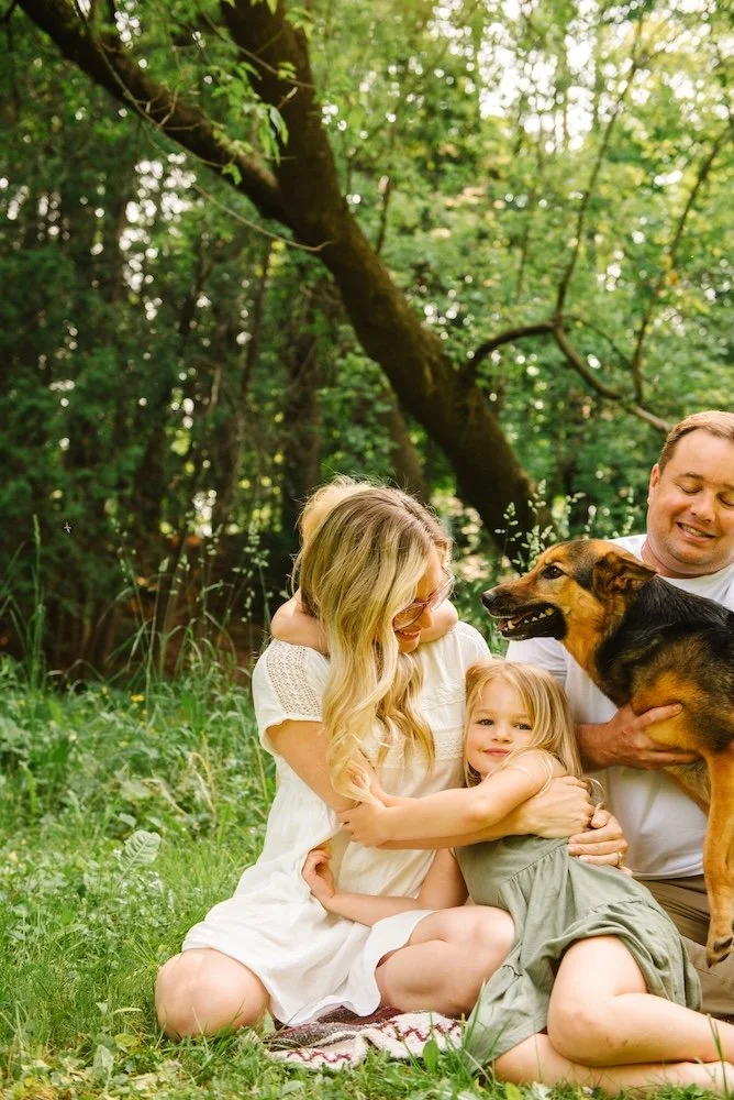 A family of three, two women and a man, sitting on a blanket on the grass in a forested area. They are hugging and smiling, with a dog in the man's arms. The scene is bright and cheerful, with trees and greenery in the background.
