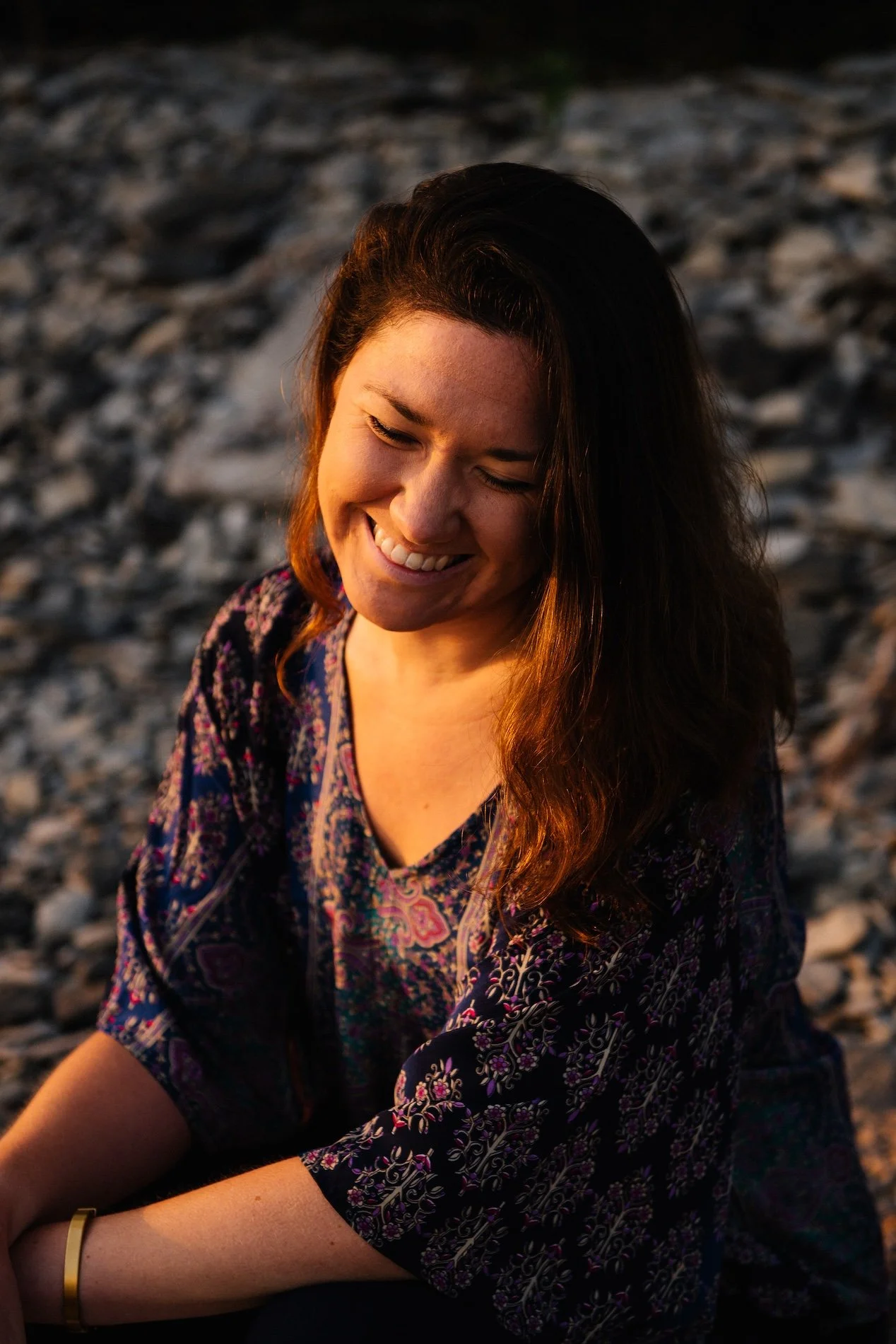 A woman with long, wavy hair is smiling and looking downward, sitting on a rocky surface during sunset.