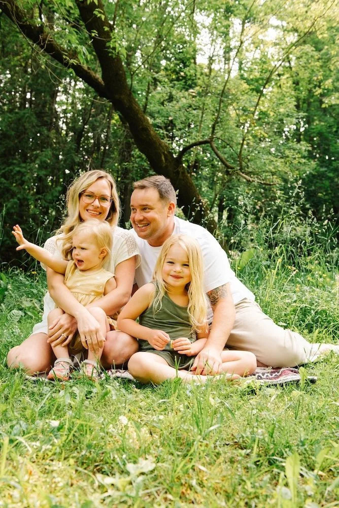 Family of four sitting on a blanket in a lush green forest, smiling and enjoying outdoor time. The parents have blonde hair, and the children are young girls, one with blonde hair, and one with light-colored hair, all dressed casually.