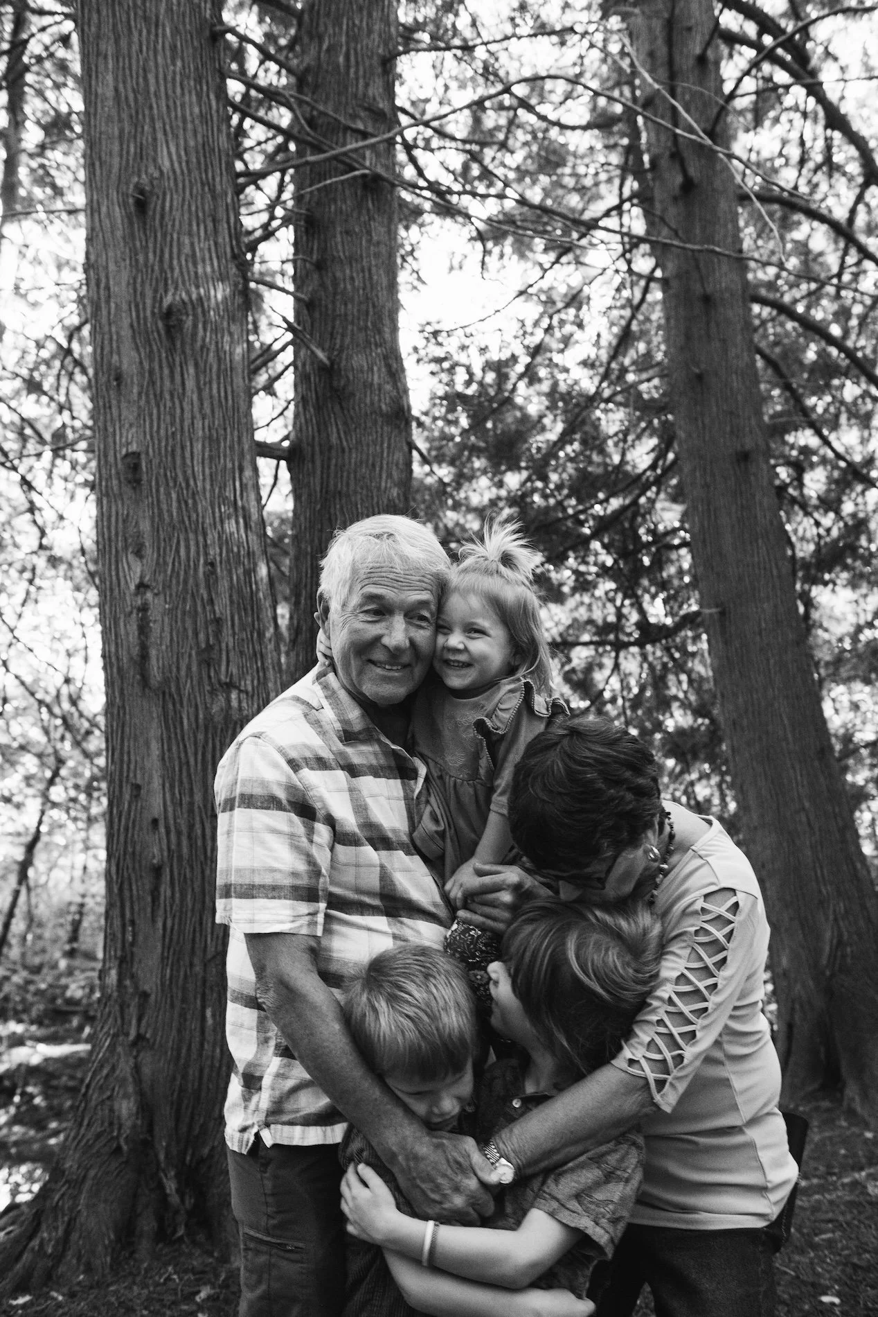 A multigenerational family of five in a forest, embracing and smiling, black and white photograph.
