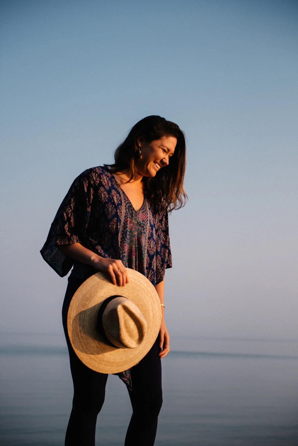 Woman standing outdoors near water, holding a straw sun hat, smiling, during sunset or sunrise.