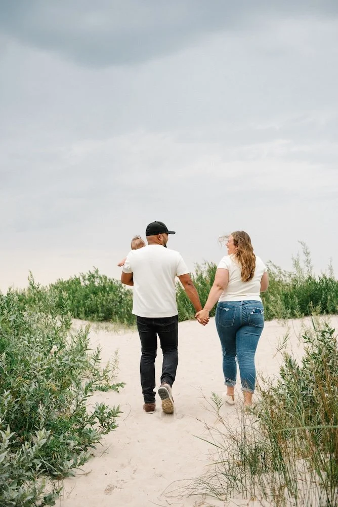 A family walking hand in hand on a sandy beach path with wind-blown hair, surrounded by green bushes and under a cloudy sky.