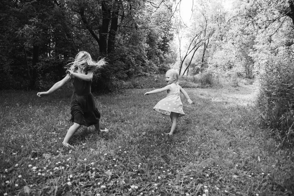 Two young girls playing and dancing in a grassy field surrounded by trees, one with long hair and the other with short hair, wearing dresses, in a nature setting.