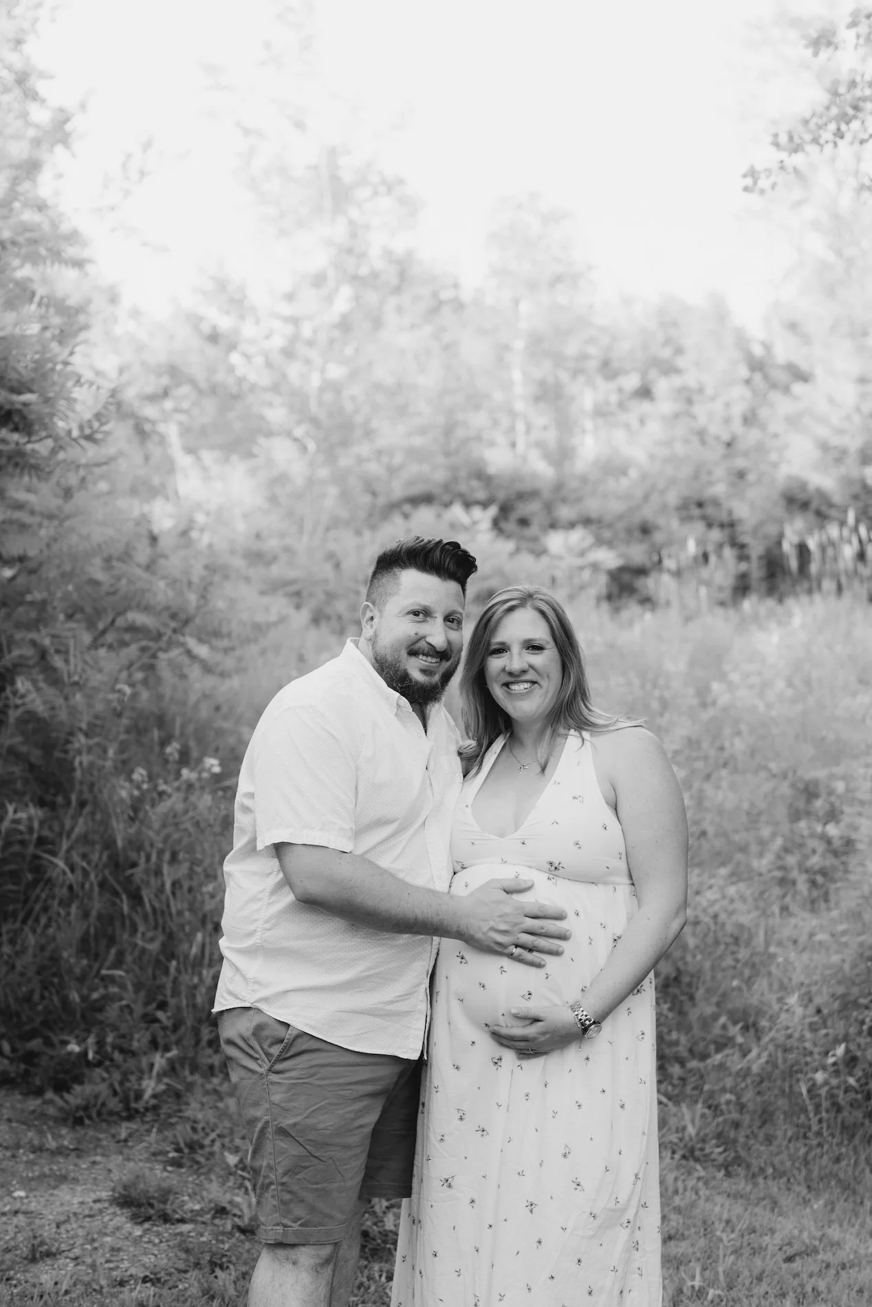 A man and pregnant woman smiling outdoors in a natural setting, both resting their hands on her baby bump.