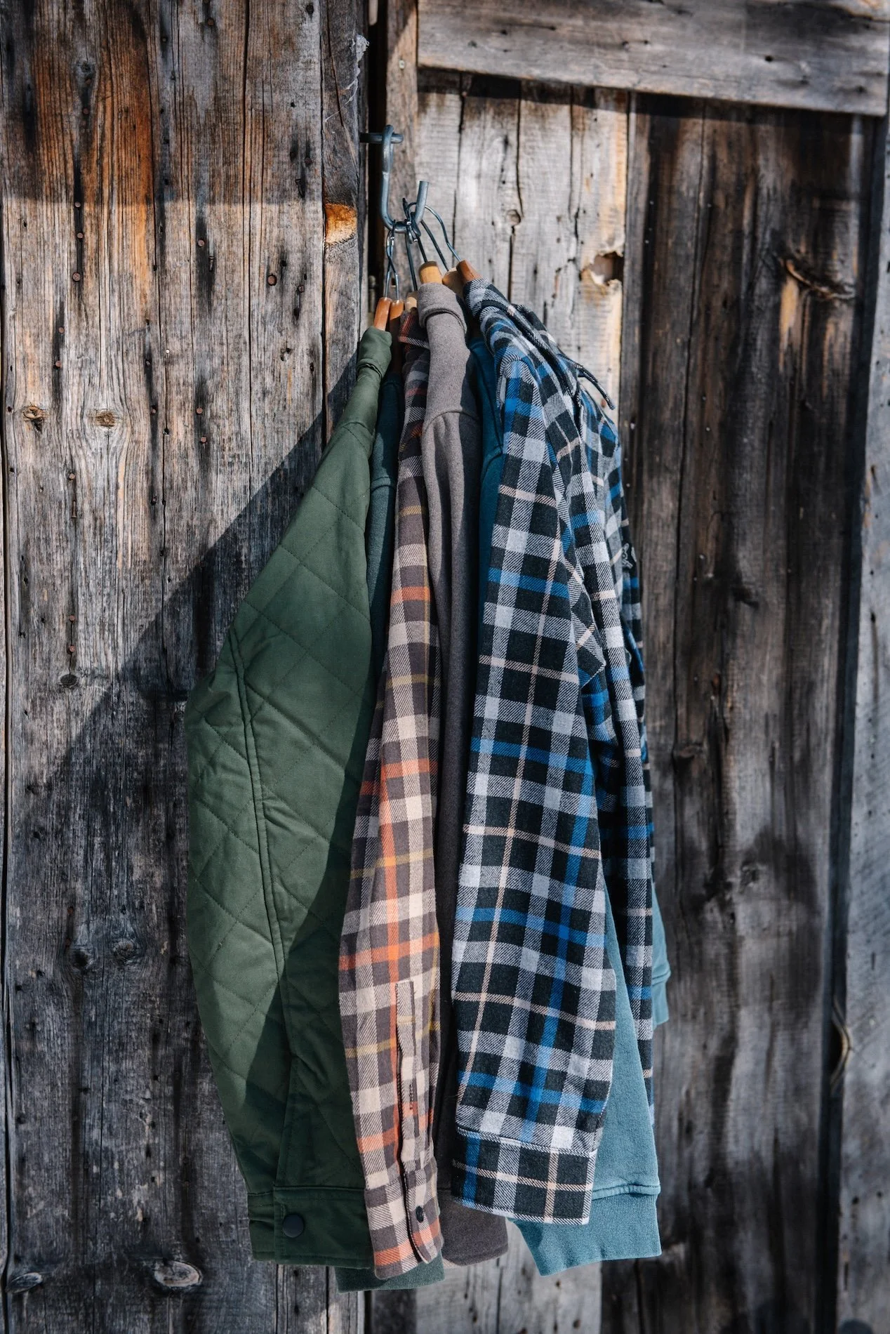 A set of four plaid and solid-colored shirts hanging on a hook on a weathered wooden wall.
