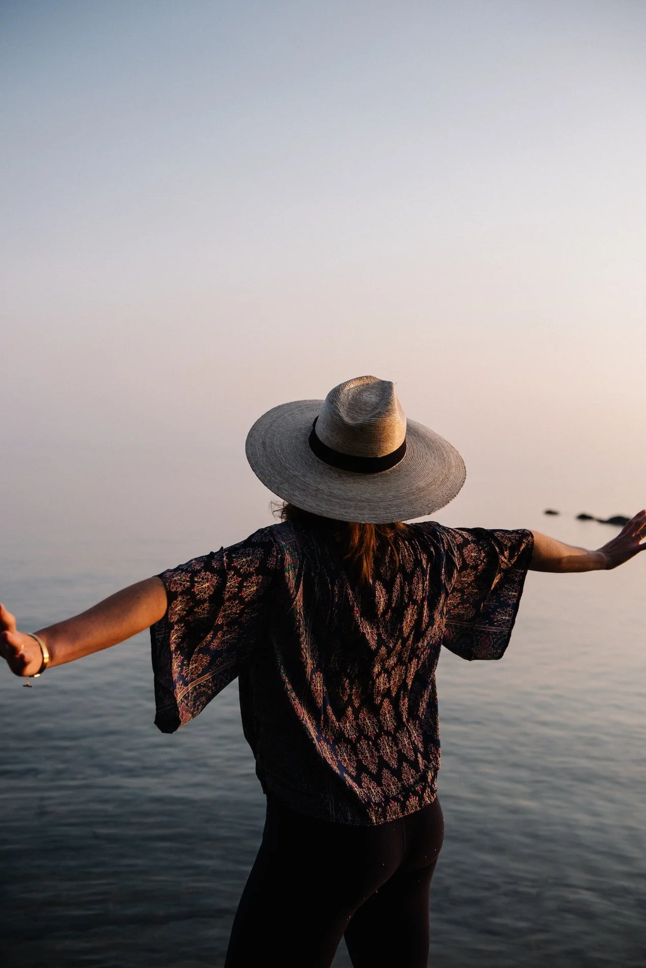 A woman with arms outstretched by the water at sunset, wearing a large sun hat and patterned top.