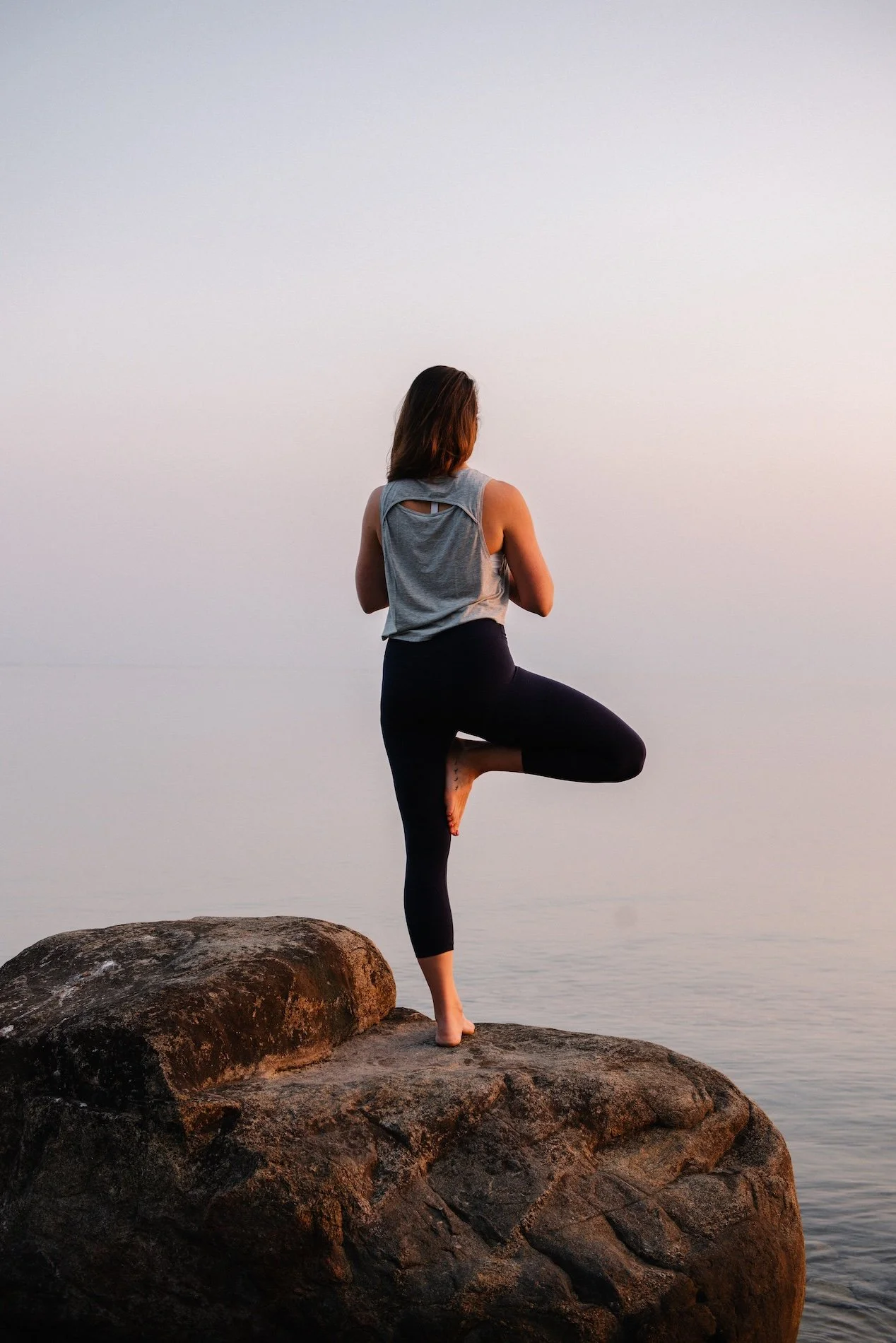A woman practicing yoga on a large rock by the water during sunset, standing on one leg with the other foot resting on her inner thigh, in a tree pose.