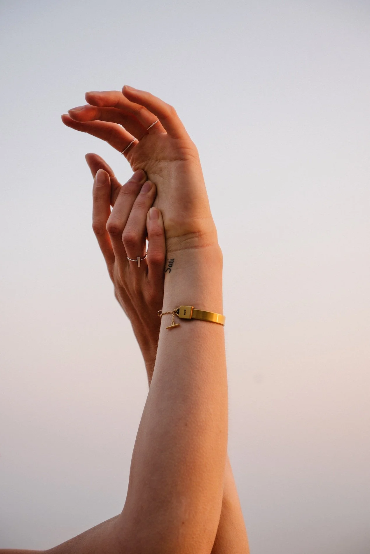 Two hands are intertwined against a plain light-colored sky. One hand appears to be supporting the other, with fingers gently wrapped around the wrist and palm. The person wearing the supporting hand has a gold bracelet and a thin silver ring, with a small tattoo on the wrist that reads 'Salt'.