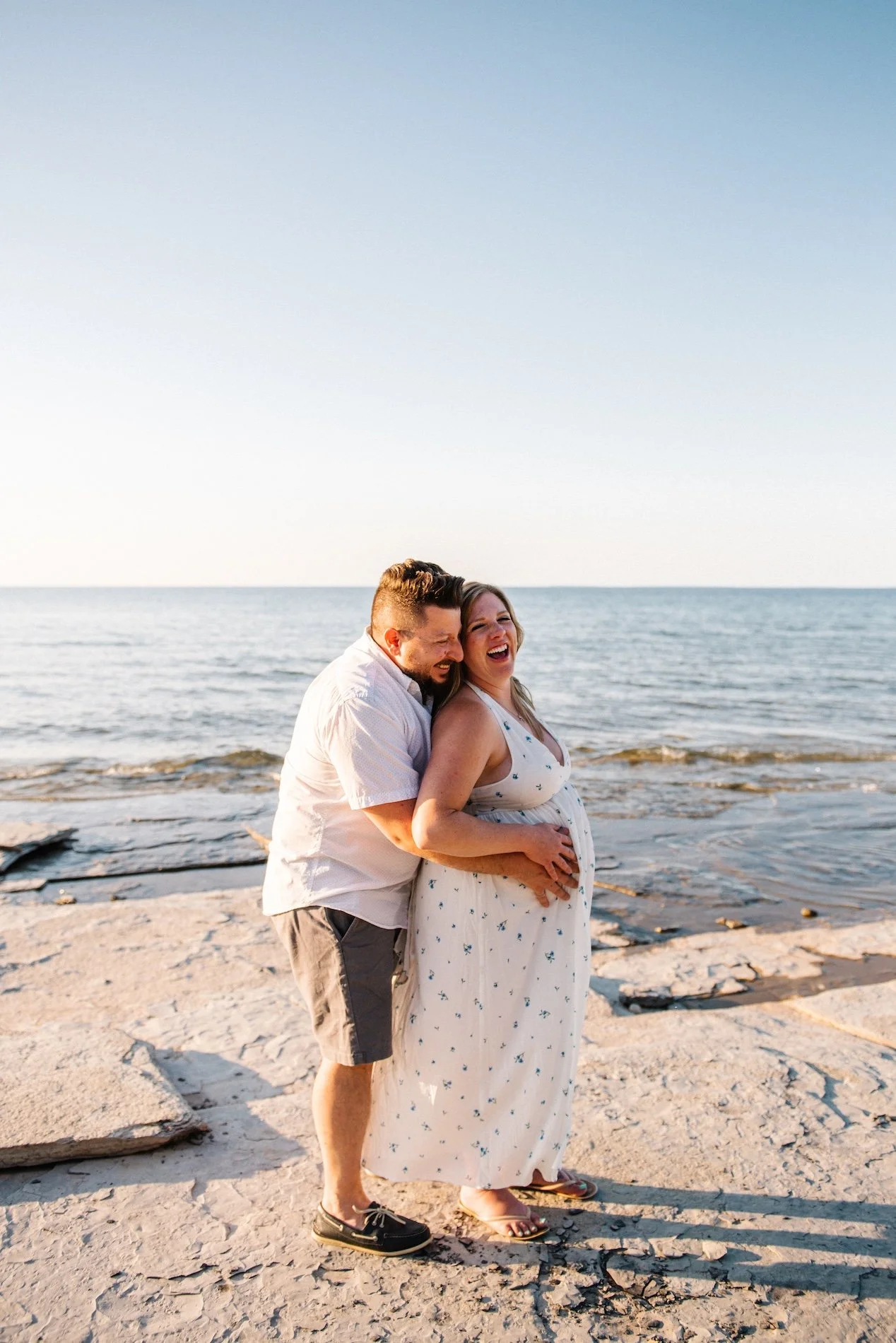 A smiling pregnant woman in a white dress with a blue floral pattern standing on a rocky beach with a man hugging her from behind, both enjoying the sunset by the water.