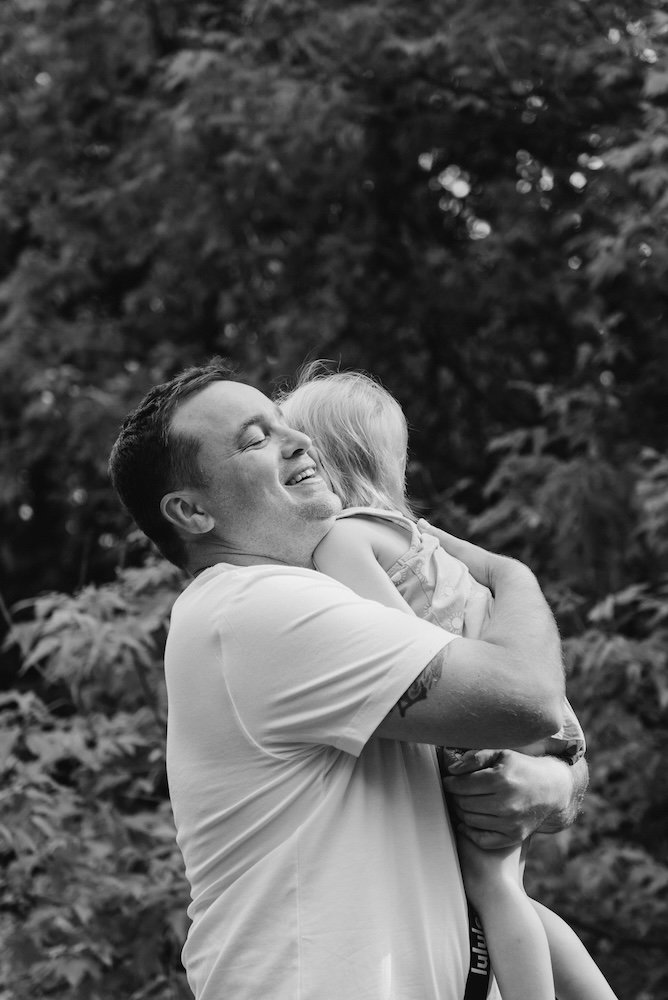 A man holding a young girl in an outdoor setting with trees in the background, smiling and embracing her.