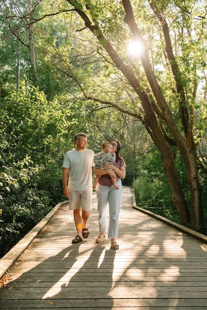 A family of three walking on a wooden boardwalk through a lush green forest, with sunlight shining through the trees.