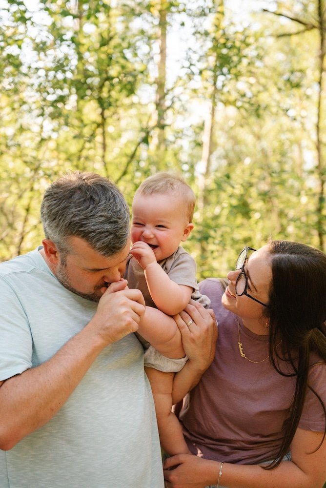 Family of three enjoying a moment outdoors: a man holding a laughing baby girl, a woman wearing glasses looking at them, surrounded by green trees in a park.