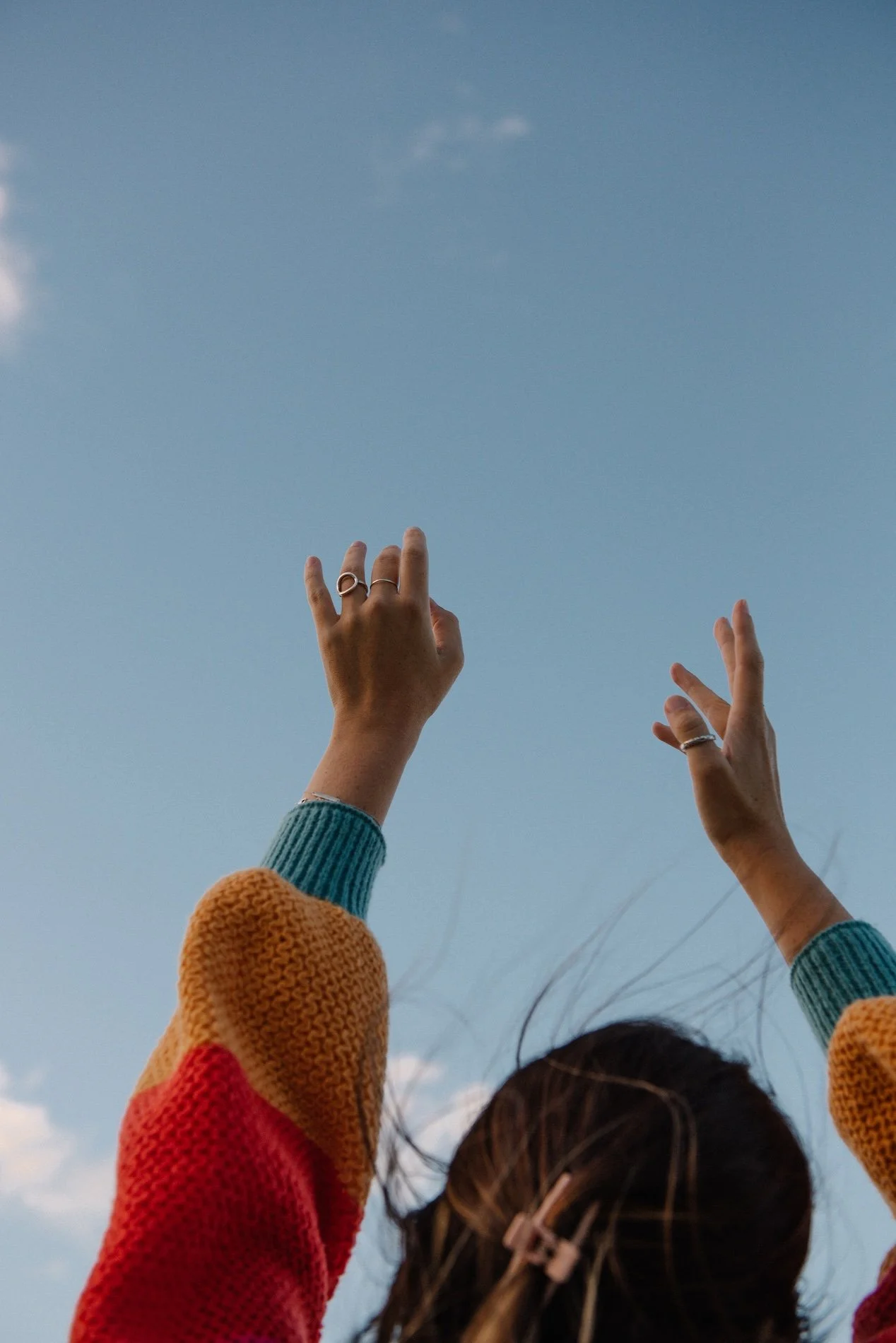 A person with long hair and colorful clothing reaches up towards a clear blue sky with a few small clouds.