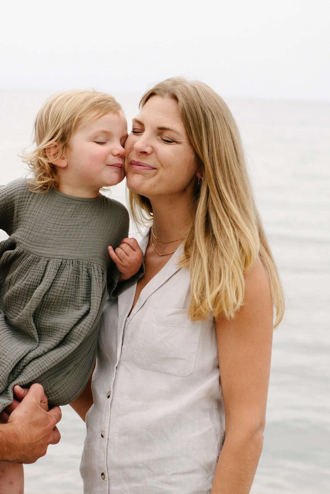 A woman and a young girl sharing a tender moment near a body of water. The girl is kissing the woman's cheek, and both appear to be enjoying the moment. The woman has blonde hair and is wearing a sleeveless white shirt, while the girl has curly blond