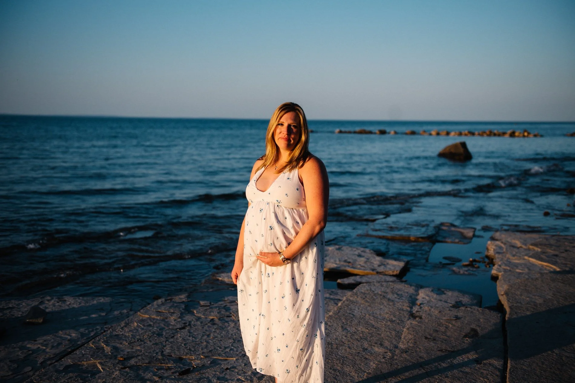 Woman in a white dress with small floral patterns standing on a rocky beach at sunset, with the ocean and rocks in the background.