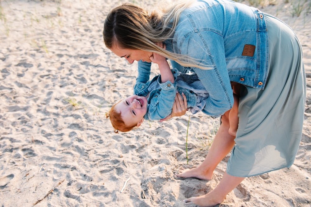 A woman and a young child playing on a sandy beach, with the woman bending over and holding the child, who is smiling and laughing.