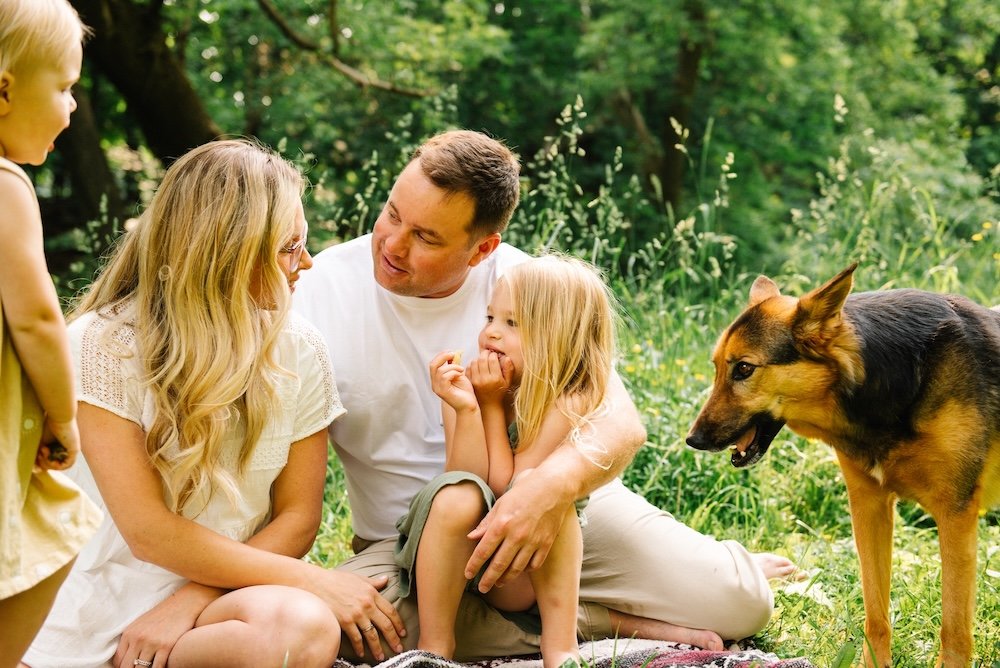 A family of four, including two children, a woman, a man, and a dog, sitting on the grass in a lush green park, enjoying each other's company.