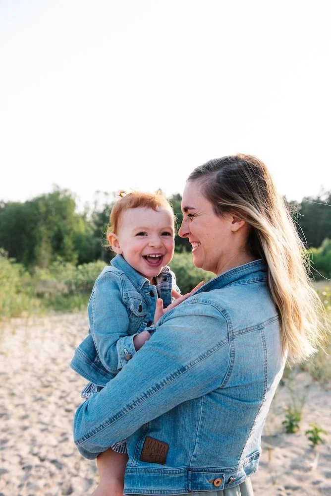 A woman holding a smiling toddler girl outdoors on a sandy area with greenery in the background.