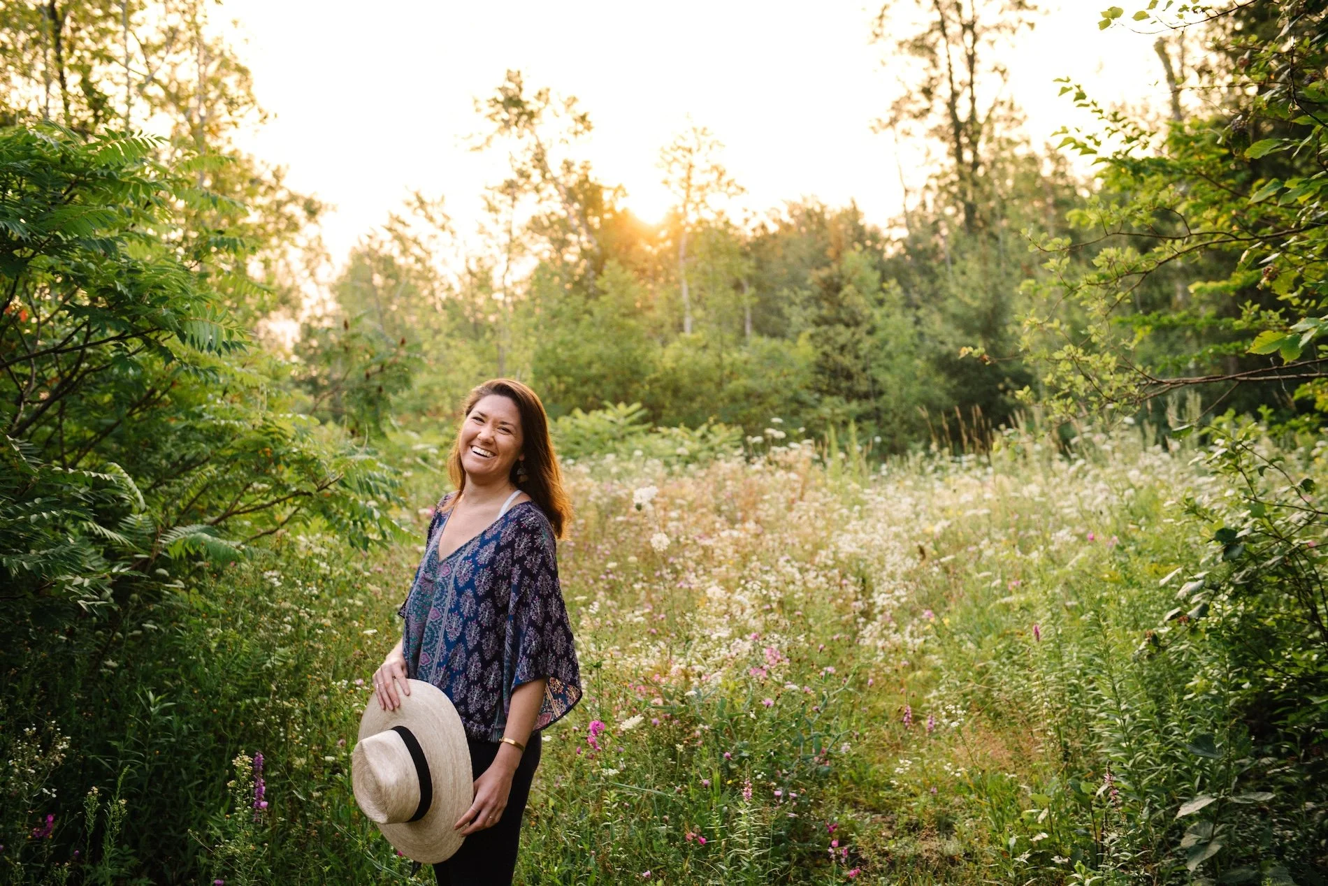 A woman smiling and holding a straw hat in a lush green field with wildflowers during sunset.