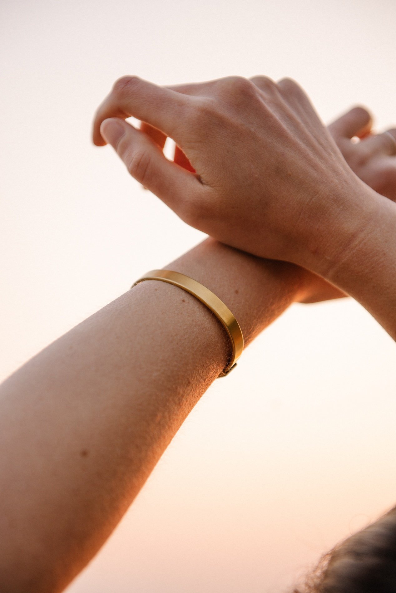 Close-up of two hands with one hand gently holding the wrist of the other, wearing a gold bracelet, against a neutral background.