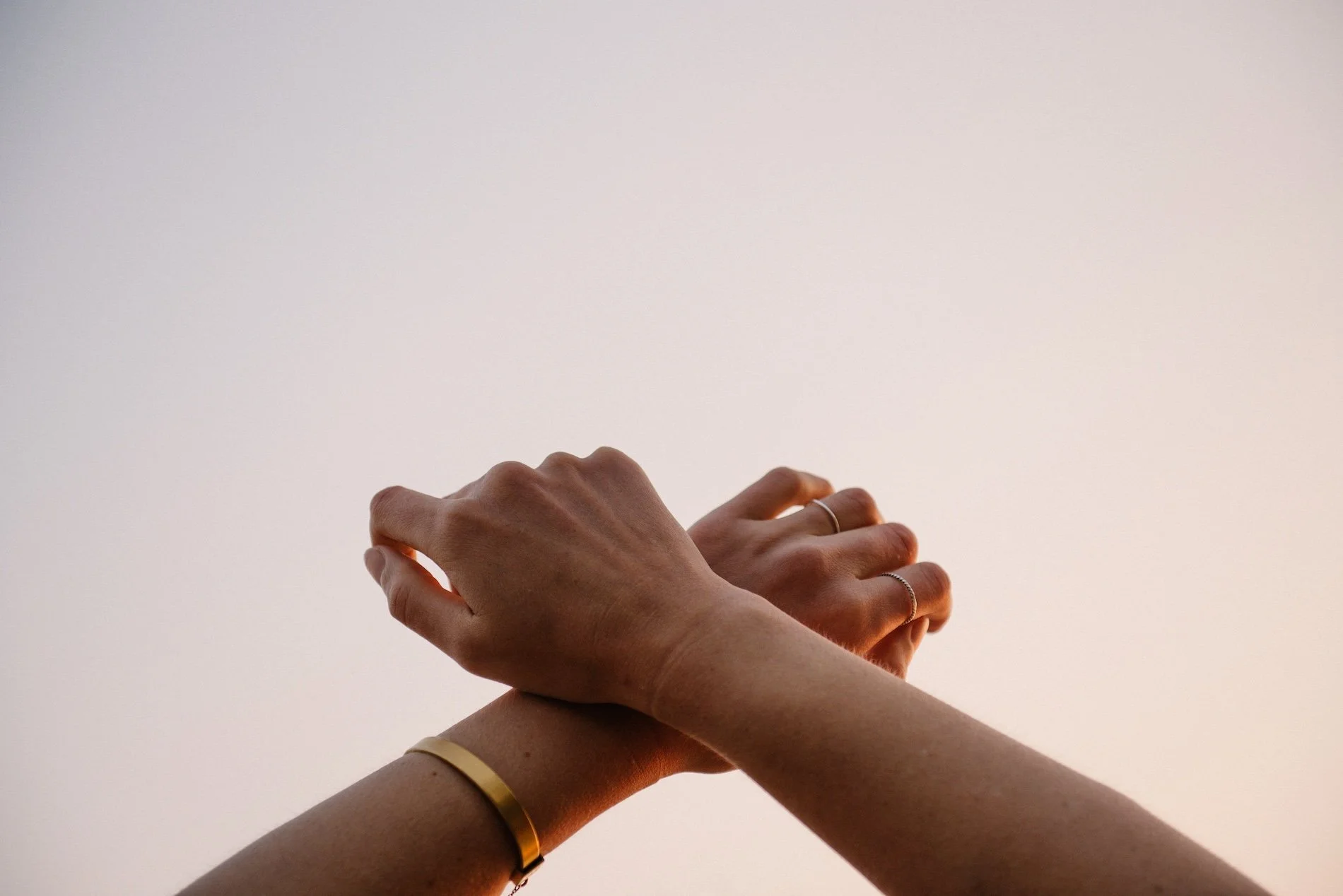 Two hands overlapping, one wearing a gold bracelet and rings, against a plain background.