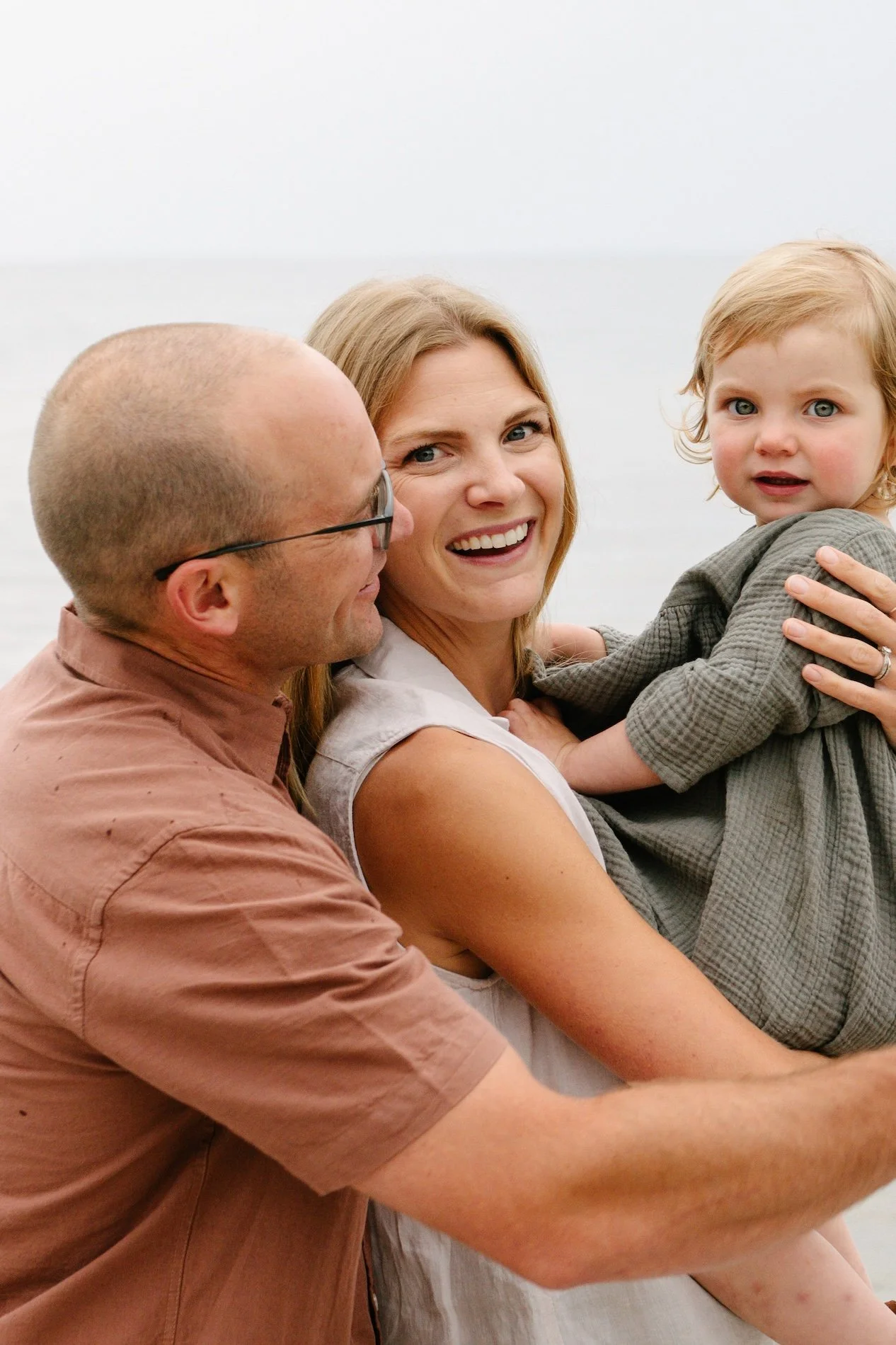 A smiling woman holding a child and a man with glasses looking at her, on a beach with the ocean in the background.
