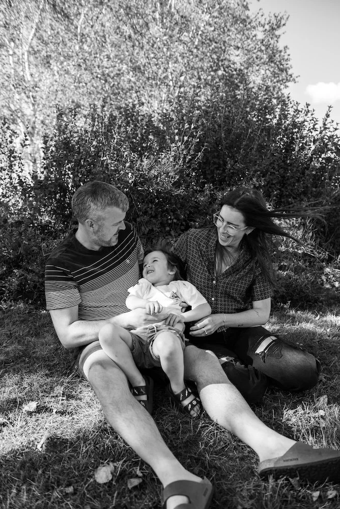 A family of three sitting on the grass outdoors, smiling at each other, with a bush and trees in the background, in a black and white photograph.