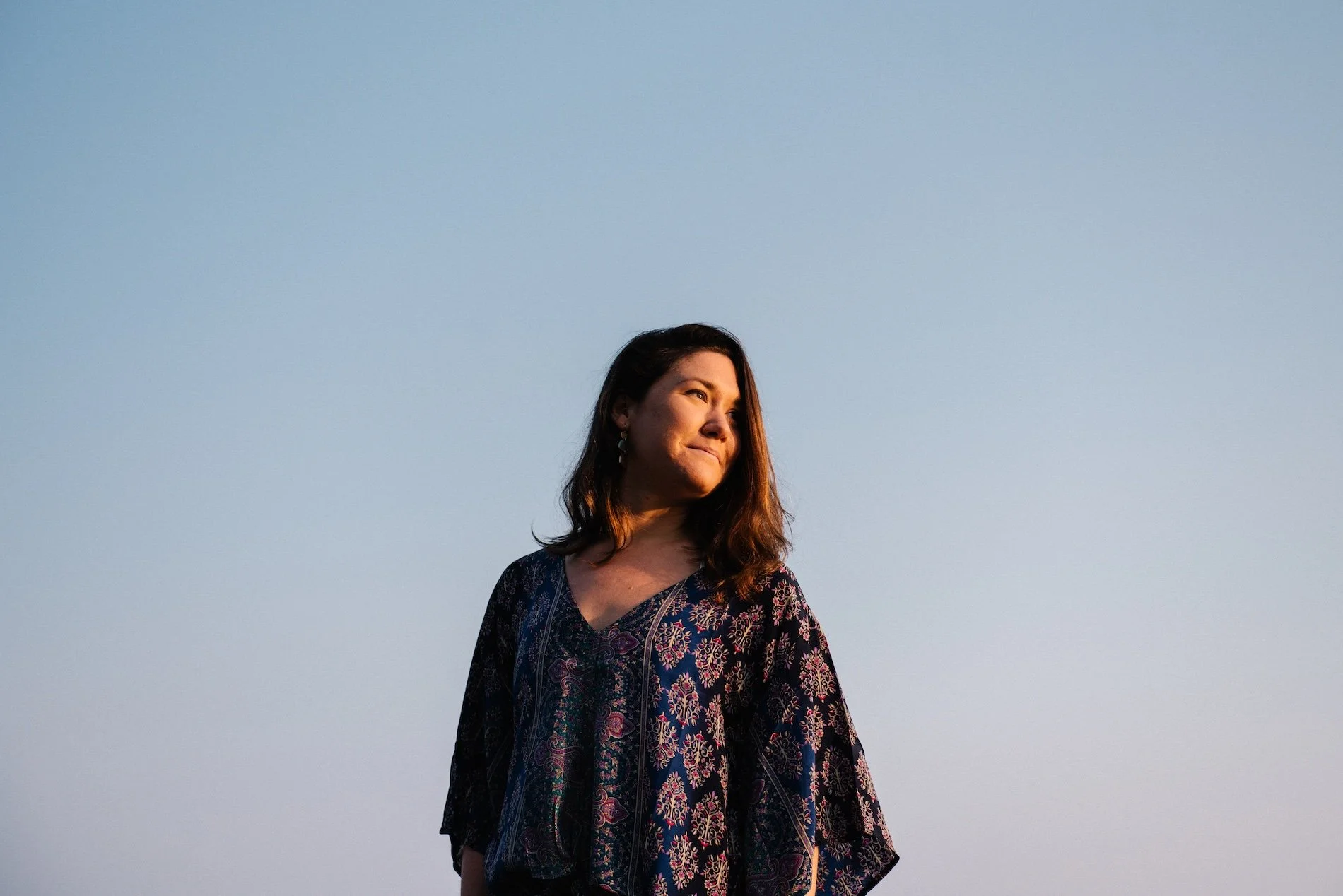 A woman with shoulder-length brown hair, wearing a patterned blouse, stands outdoors during sunset, gazing to the side with a thoughtful expression against a clear sky.