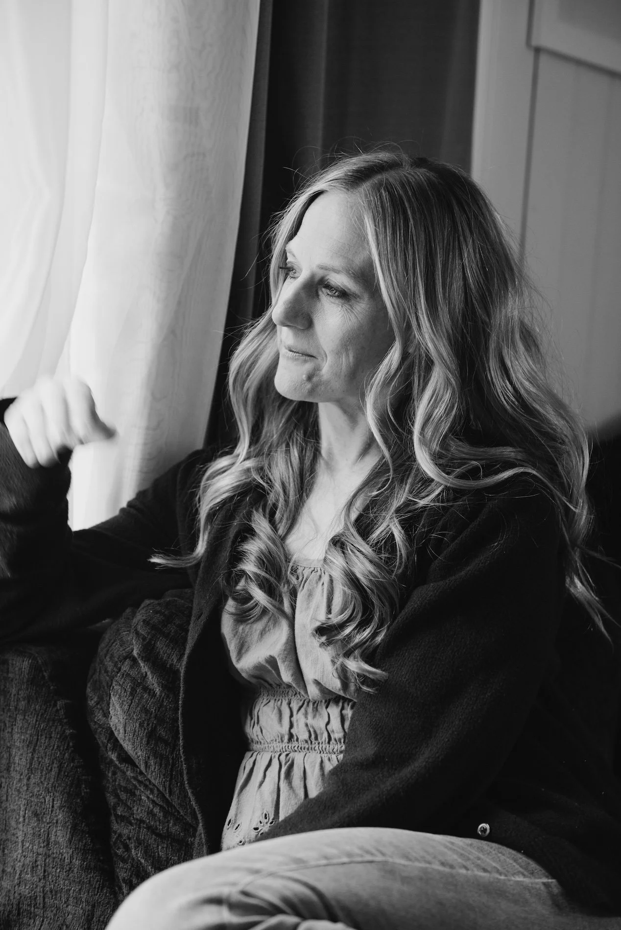 A woman with long wavy hair sitting near a window and looking outside, captured in black and white.