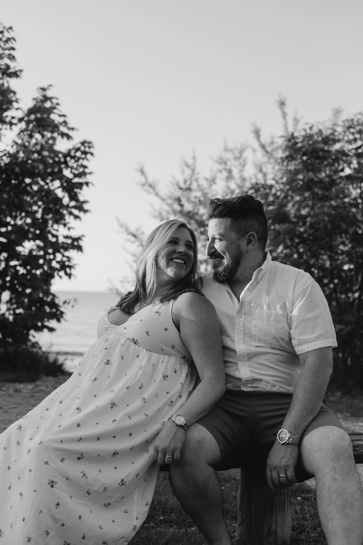 A couple sitting outdoors near a beach, smiling and looking at each other, black and white photo.