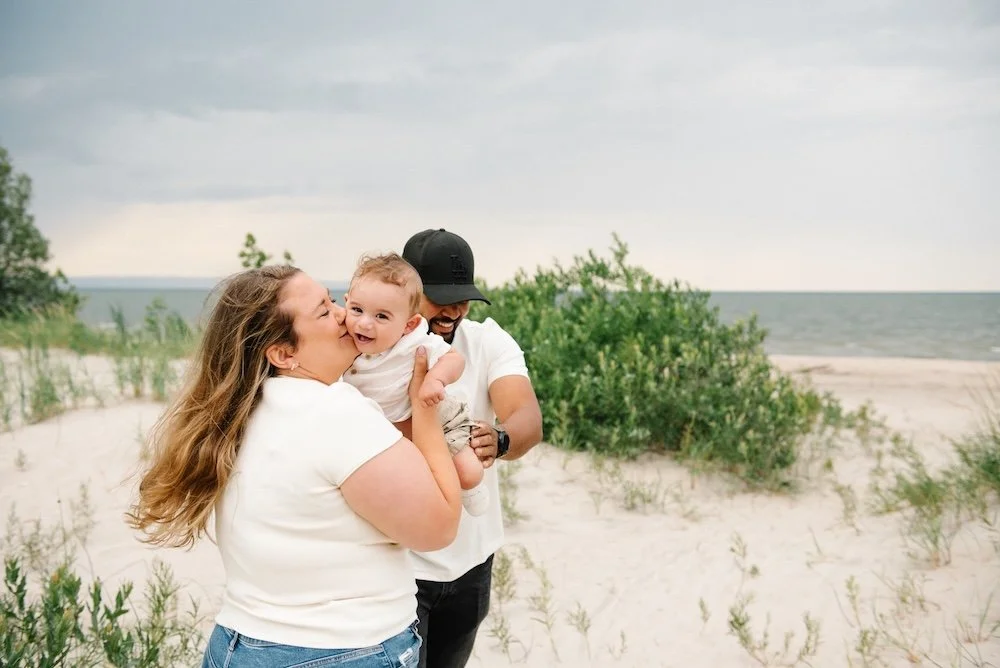 A family enjoying time on a sandy beach, with the ocean and clouds in the background. The woman is kissing a smiling baby, while the man holds the baby and laughs.